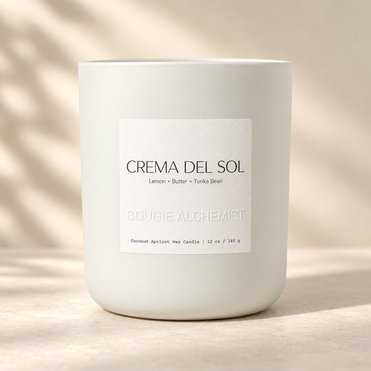 A white ceramic jar candle with a minimalist white label reading 'CREMA DEL SOL' and 'BOUGIE ALCHEMIST', set on a warm beige surface with soft leaf shadows.