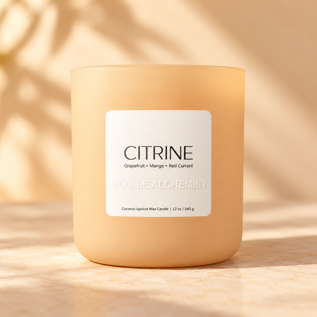 A frosted glass candle jar in a warm peachy-orange hue with a white label reading 'CITRINE' and 'BOUGIE ALCHEMIST'. The candle sits on a sunlit beige surface with soft leaf shadows cast across it.