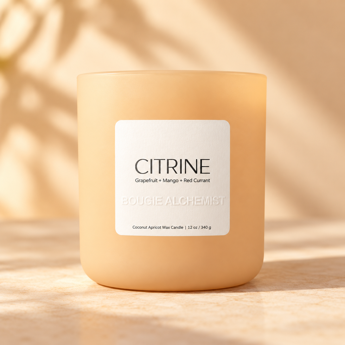 A frosted glass candle jar in a warm peachy-orange hue with a white label reading 'CITRINE' and 'BOUGIE ALCHEMIST'. The candle sits on a sunlit beige surface with soft leaf shadows cast across it.