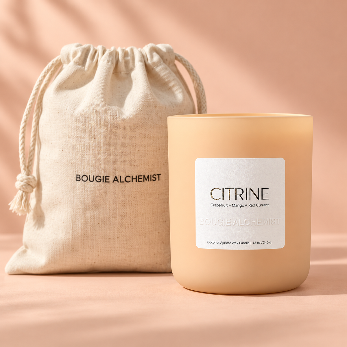 A Bougie Alchemist 'Citrine' coconut apricot wax candle in a frosted peach-colored glass jar, shown next to its matching beige linen drawstring pouch on a warm peach background.