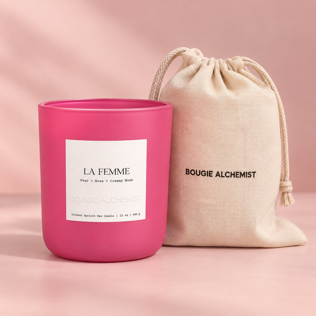 A bright pink scented candle jar labeled 'LA FEMME' stands next to a beige drawstring pouch printed with 'BOUGIE ALCHEMIST' on a soft pink background.