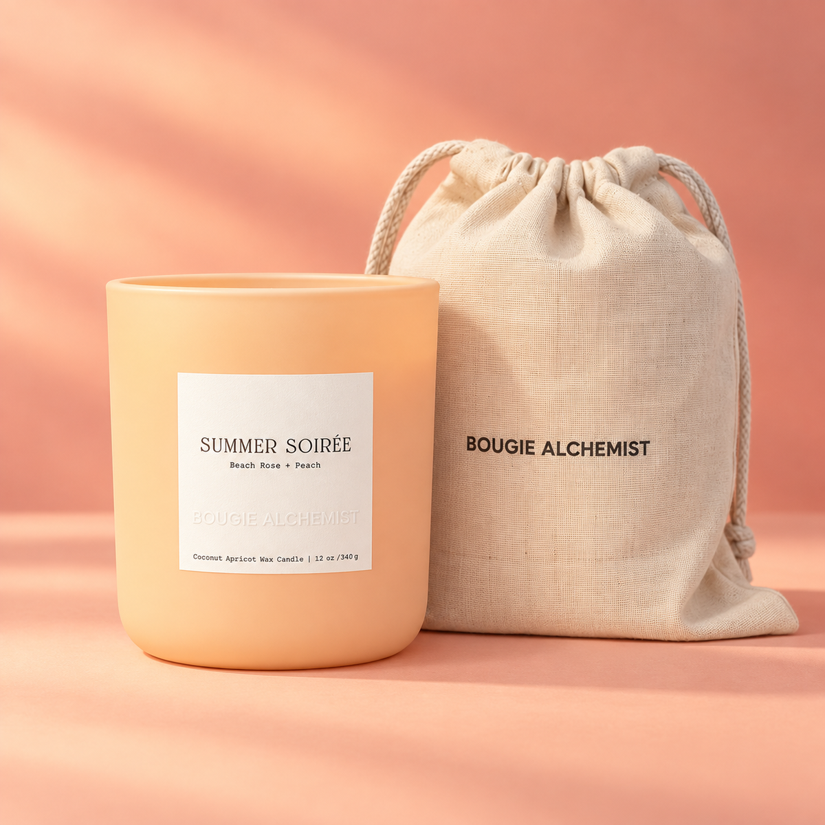 A peach-colored scented candle in a matte glass jar with a white label reading 'SUMMER SOIRÉE', standing next to a beige linen drawstring pouch printed with 'BOUGIE ALCHEMIST'. The products are set against a warm peach-pink background with soft shadows.