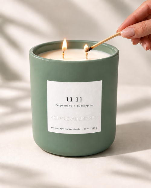 A sage green candle jar with two lit wicks and burning flames, being lit with a wooden match by a hand. The candle features a white label with the brand name 'BOUGIE ALCHEMIST' and scent '1111 Peppermint + Eucalyptus'.