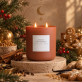 A terracotta-colored double-wick Bougie Alchemist candle labeled 'Spiced Gingerbread' sits on a stone pedestal in a cozy holiday setting with cinnamon sticks, ginger, star anise, and gingerbread cookies.