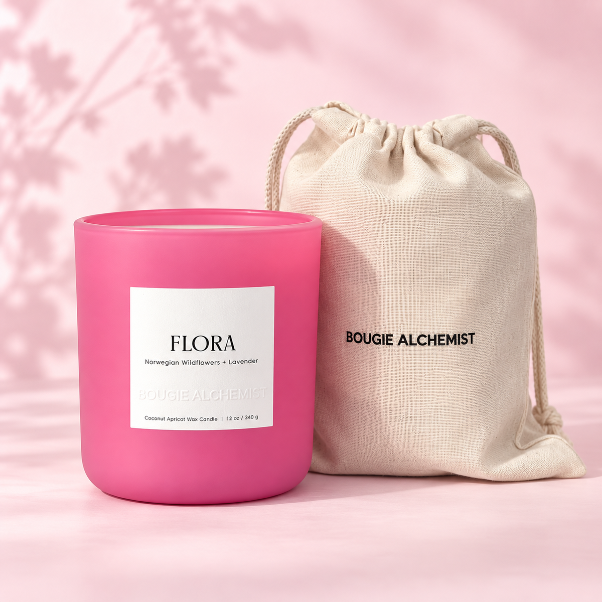 A bright pink scented wax candle jar with a white label reading FLORA, standing next to a beige drawstring pouch printed with BOUGIE ALCHEMIST, set against a soft pink background with leaf shadows.