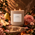 A lit double-wick candle in a matte beige jar sits on stone steps surrounded by pink flowers, a pear, rose petals, and warm decorative elements. The front label reads 'LUNA' with smaller text 'Pine + Cherry Blossom + Dark Musk' and 'BOUGIE ALCHEMIST.