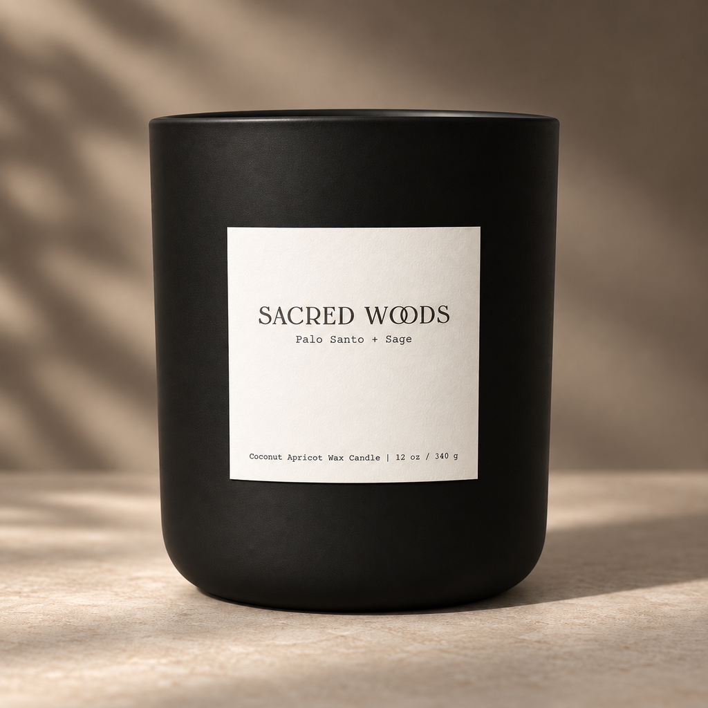 A matte black Bougie Alchemist candle jar with a white rectangular label reading 'SACRED WOODS' and 'Palo Santo + Sage' on a warm beige surface with soft shadow overlays.