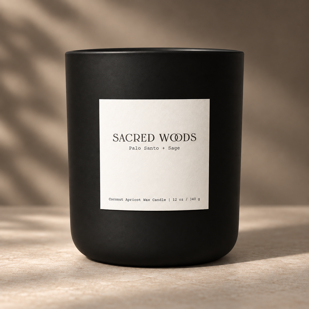 A matte black Bougie Alchemist candle jar with a white rectangular label reading 'SACRED WOODS' and 'Palo Santo + Sage' on a warm beige surface with soft shadow overlays.
