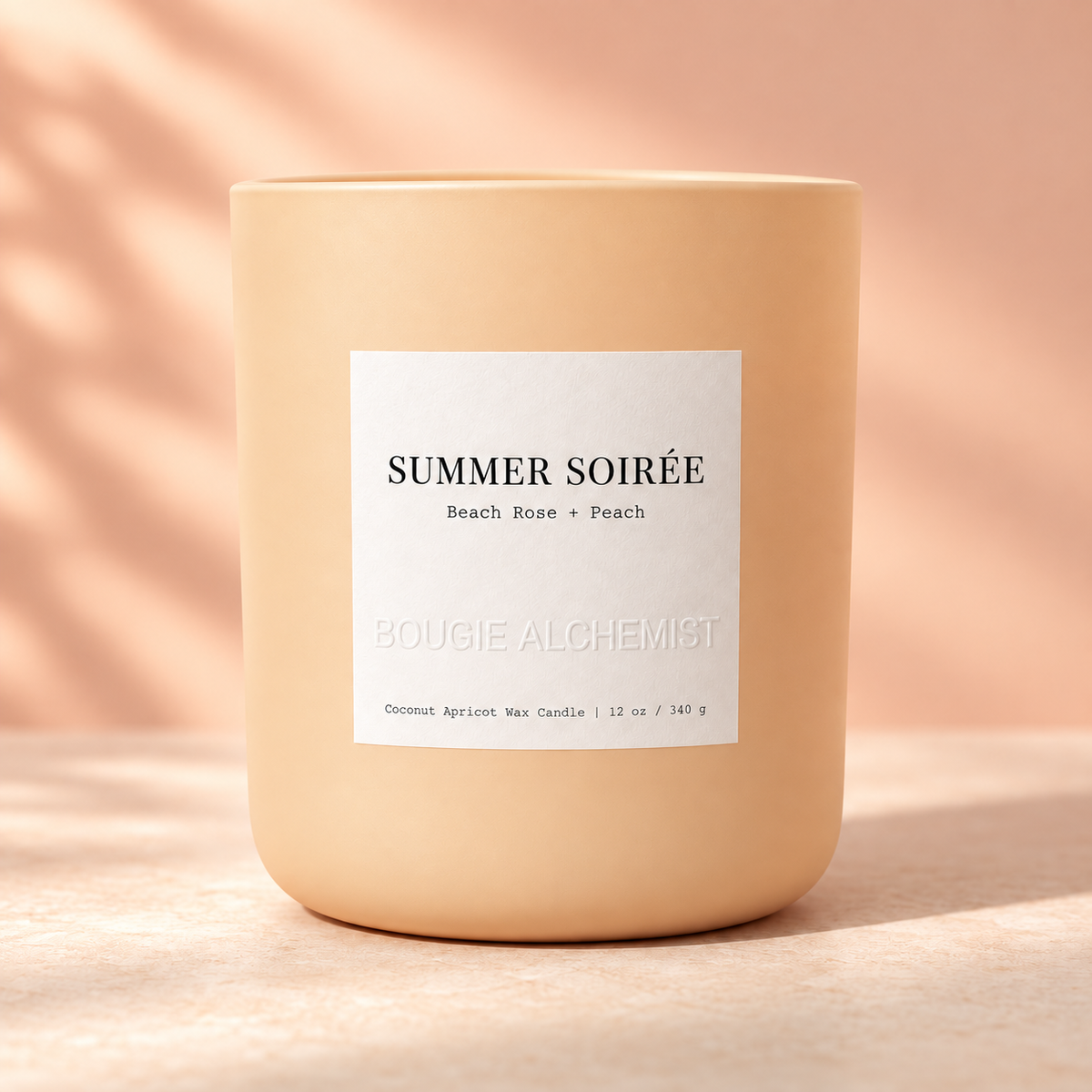 A matte peach-colored candle jar with a white rectangular label reading 'SUMMER SOIRÉE Beach Rose + Peach' and 'BOUGIE ALCHEMIST', photographed on a warm beige surface with soft shadow overlays.