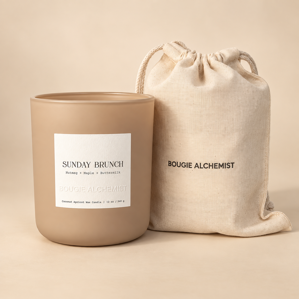 A matte taupe candle jar labeled 'SUNDAY BRUNCH' by Bougie Alchemist stands next to a matching beige drawstring pouch with the brand name printed on it, set against a warm beige background.