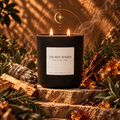 A black double-wick Bougie Alchemist candle labeled 'Sacred Woods' sits on stone steps surrounded by wood, evergreen branches, crystals, and a smudge bundle in a warm, atmospheric setting.