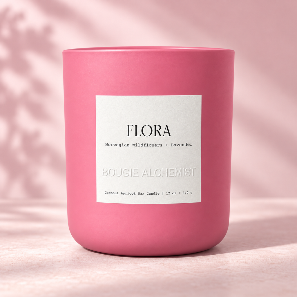 A pink ceramic candle jar with a white label reading 'FLORA' and 'BOUGIE ALCHEMIST', photographed on a soft pink surface with a matching pink background and subtle leaf shadows.