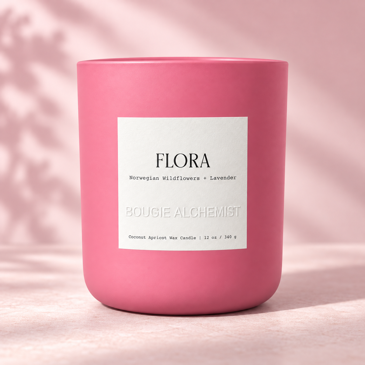 A pink ceramic candle jar with a white label reading 'FLORA' and 'BOUGIE ALCHEMIST', photographed on a soft pink surface with a matching pink background and subtle leaf shadows.