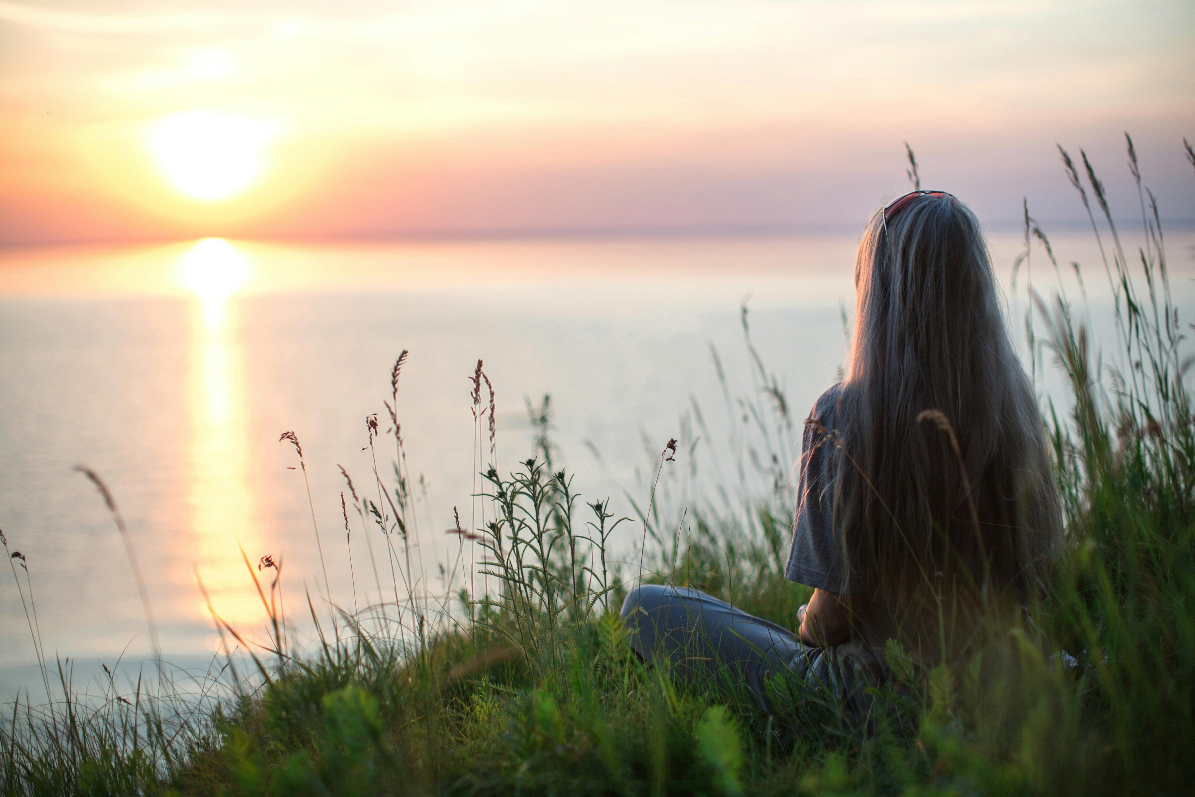 Person sitting in a field of tall grass watching a sunset over water.